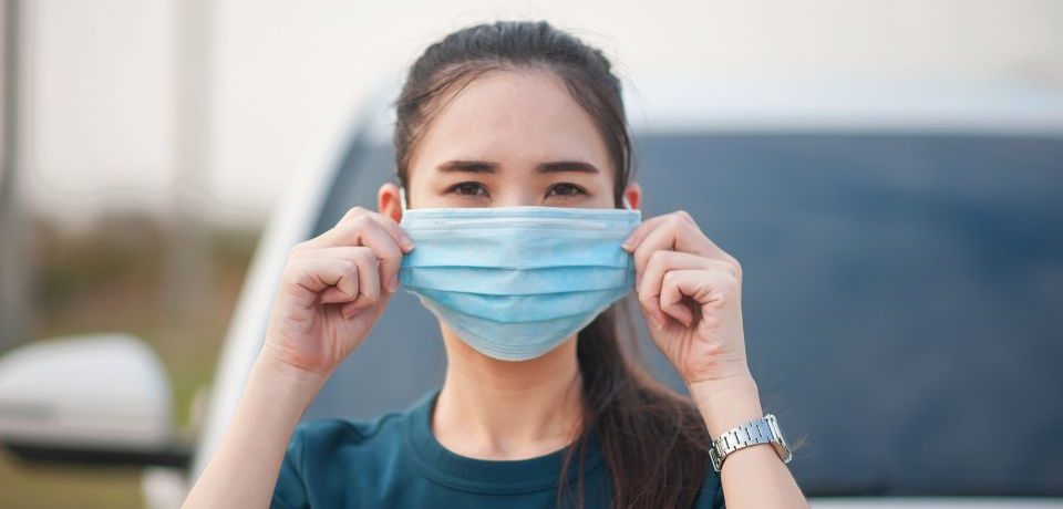 A woman outdoors putting on a surgical face mask.