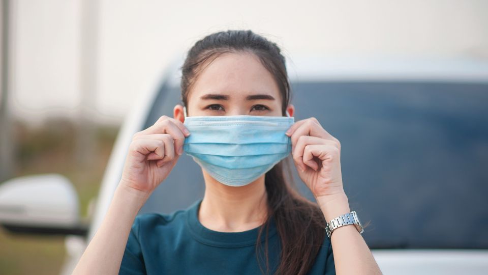 A woman outdoors putting on a surgical face mask.
